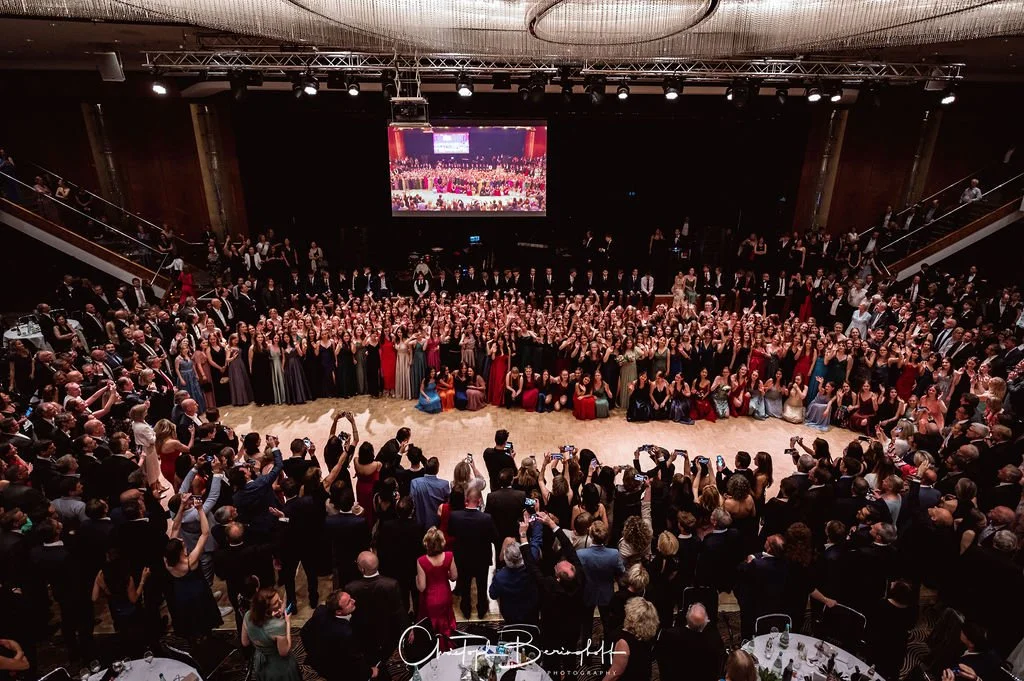 Eine große Gruppe von Menschen in eleganten Kleidern und Anzügen versammelt sich in einem großen Ballsaal für eine fotografierte Veranstaltung, mit vielen Gästen, die Fotos machen.