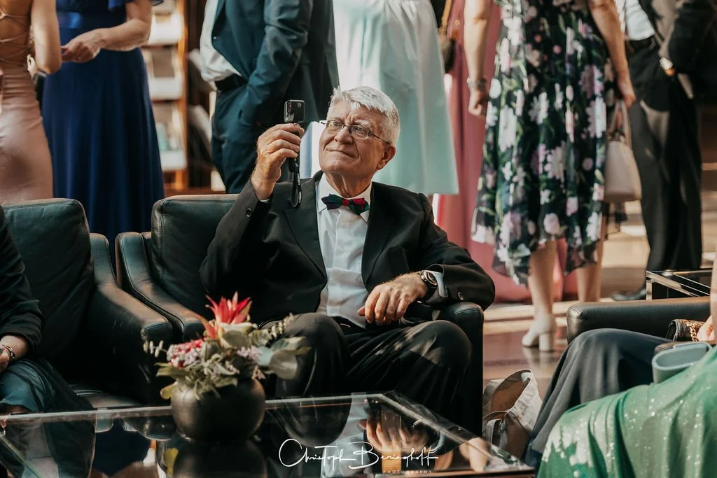 Ein älterer Mann in schwarzem Anzug mit Fliege sitzt auf einer schwarzen Ledercouch und fotografiert mit seinem Handy. Er ist in einer Gesellschaft bei einer Feier oder Veranstaltung, im Hintergrund sind mehrere Frauen in bunten Kleidern zu sehen, da