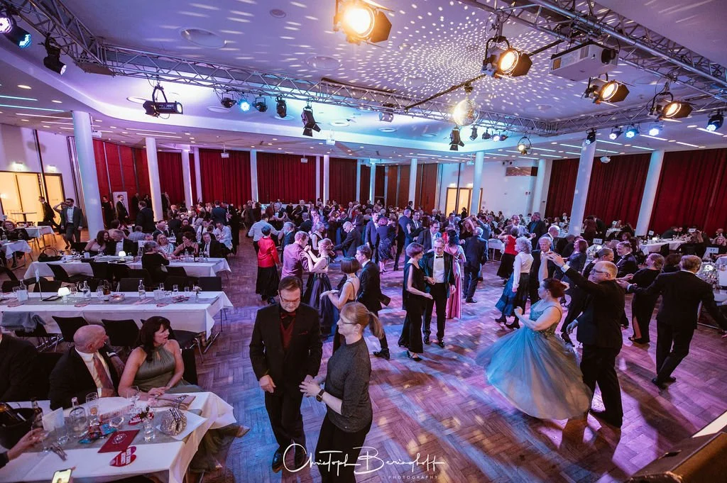Menschen tanzen auf einer großen festlichen Gala in einem Ballsaal mit rotem Vorhang, Dimmslicht und tanzender Menge.