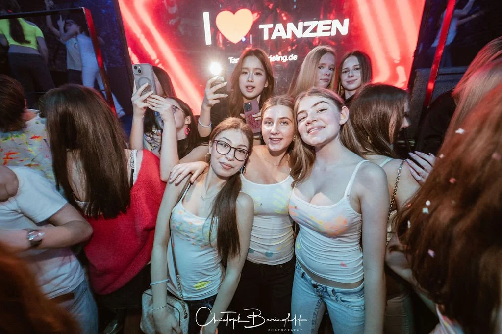 A group of young people partying at a dance event. In the background, an LED screen displays the message 'I ❤️ DANCING'.
