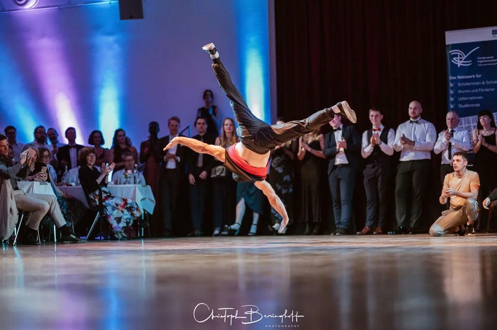 Ein Breakdancer macht eine akrobatische Bewegung auf einer Bühne vor einem Publikum, bei einer Tanzveranstaltung.