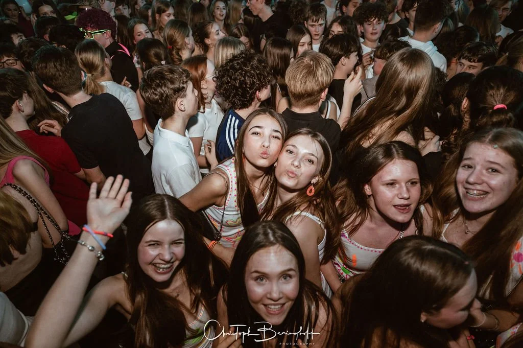 A group of young people laughing and posing at a party, with many girls in the foreground, colorful clothes, and a friendly atmosphere.