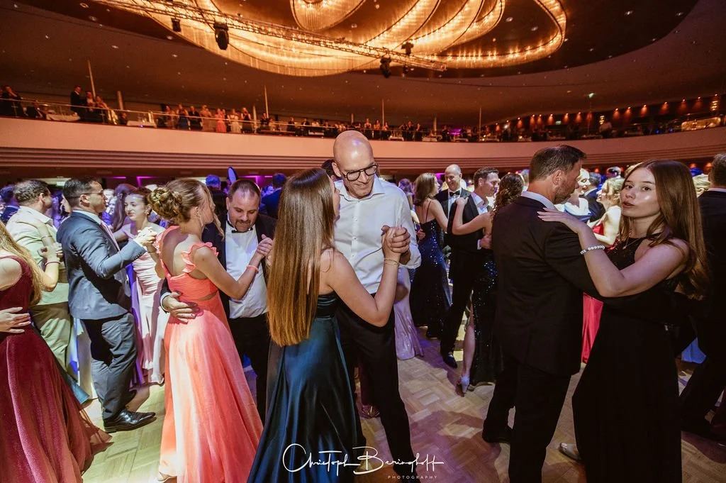 Menschen tanzen auf einer eleganten Tanzparty im Ballsaal, viele sind in festlichen Kleidern und Anzügen gekleidet, im Hintergrund eine glitzernde Decke und ein Balkon mit Zuschauern.