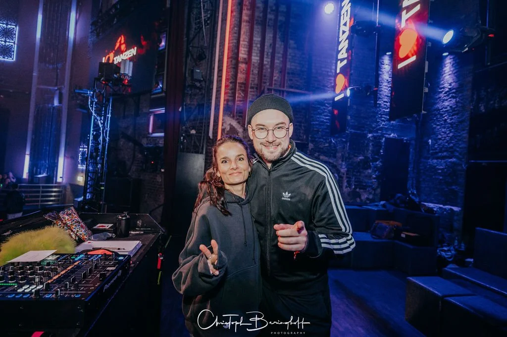 Two people in a disco or club setting—a woman and a man—smiling at the camera. There is DJ equipment in the foreground, colorful lights, and a stone-like wall in the background.