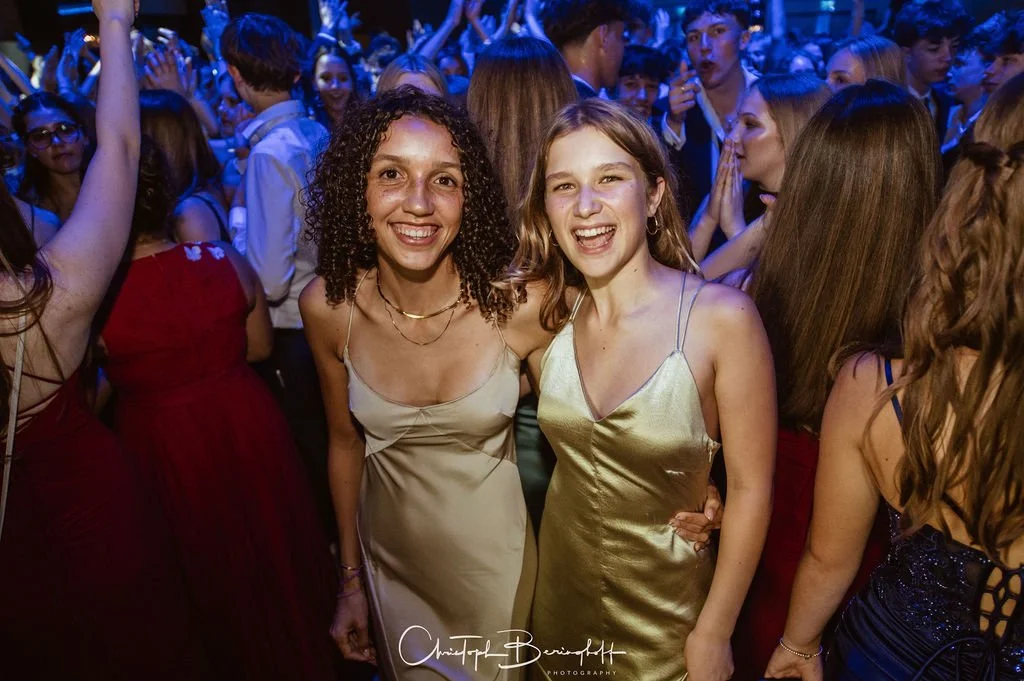Zwei junge Frauen feiern auf einer Party, umgeben von anderen feiernden Menschen im Hintergrund.