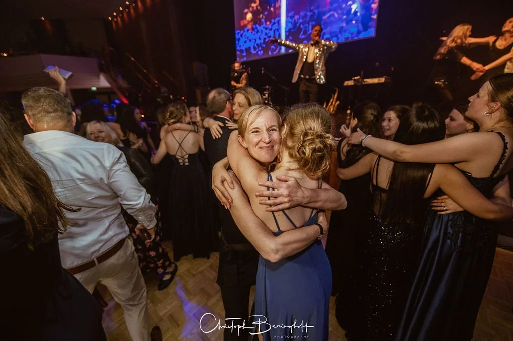 Menschen umarmen sich tanzend auf einer Party, im Hintergrund eine Bühne mit Musiker und eine große Leinwand mit digitaler Projektion.