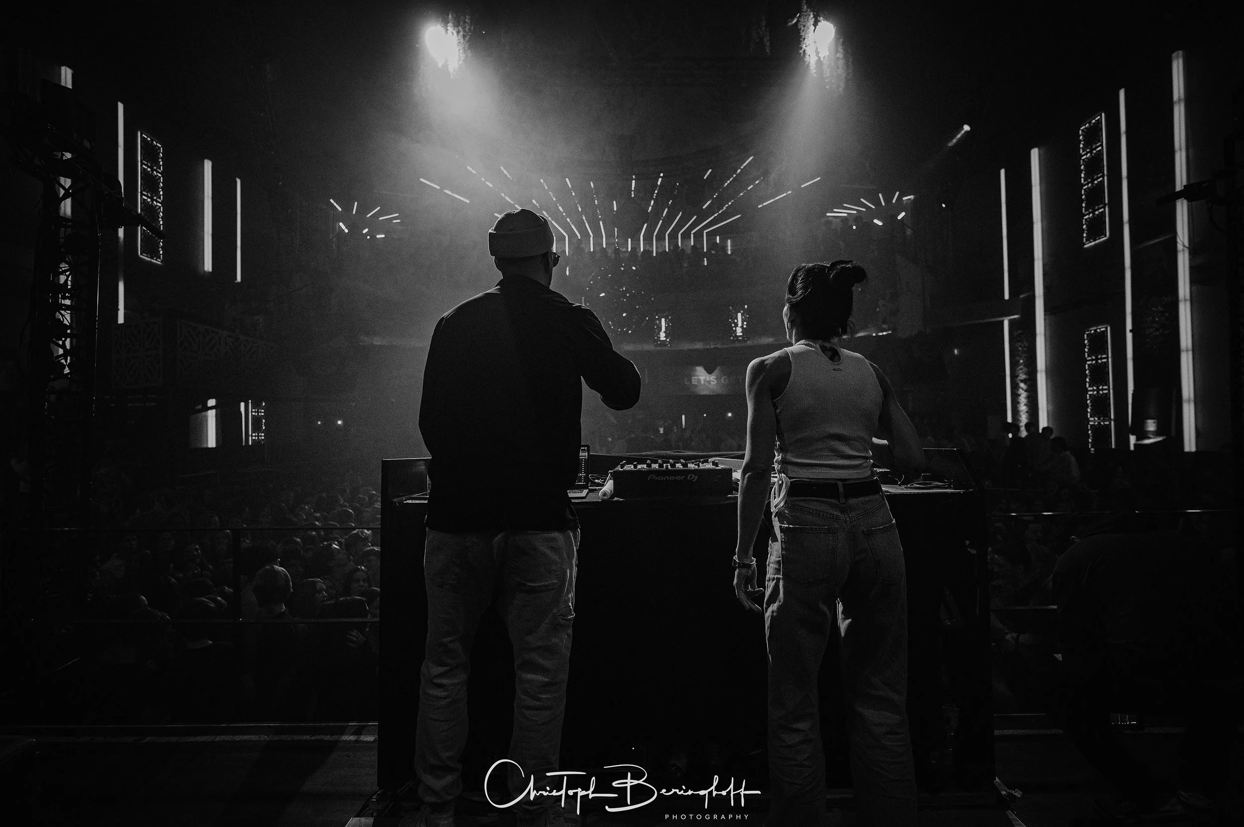 Zwei DJs stehen auf einer Bühne in einer dunklen, voll beleuchteten Club- oder Konzerthalle, während im Hintergrund eine große Menschenmenge tanzt und die Lichter und visuelle Effekte auf der Bühne lebhaft sind.