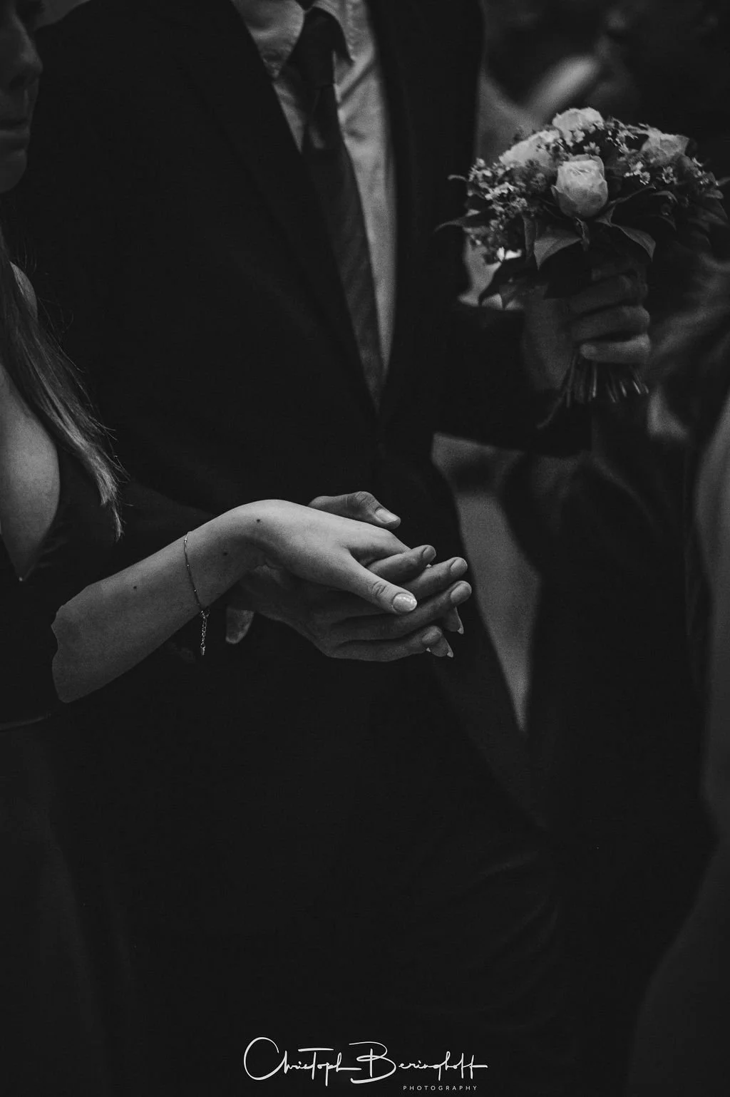 Nahaufnahme von einem Brautpaar bei einer Hochzeit, bei der der Bräutigam einen Blumenstrauß hält und die Hände der Braut und des Bräutigams sich berühren, in schwarz-weiß Fotografie.
