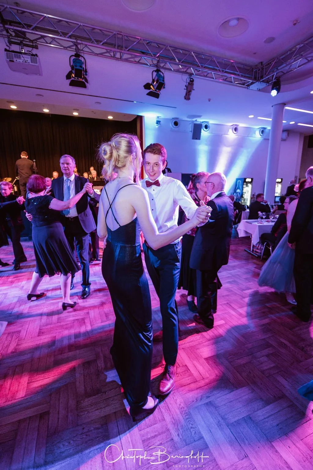 Paar tanzt auf einer eleganten Party im Saal mit anderen Gästen, die im Hintergrund ebenfalls tanzen und sitzen.