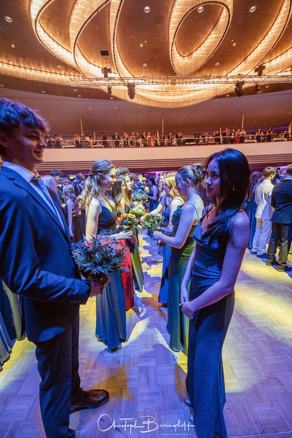 Eine Gruppe junger Menschen, hauptsächlich Frauen in eleganten Abendkleidern und Männer in Anzügen, stehen in einer Reihe im festlich beleuchteten Saal und tauschen Blumen aus, vermutlich bei einer Abschlussfeier oder Gala.