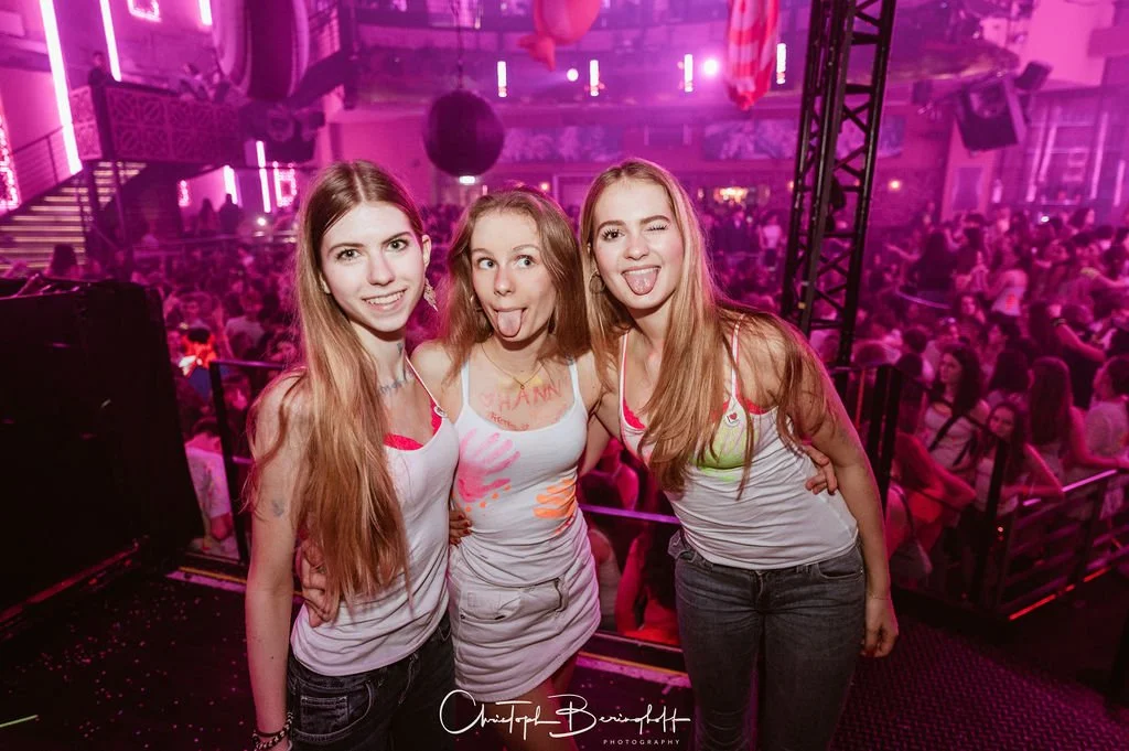 Three young women in white tank tops having fun at a party in a club with pink lighting.