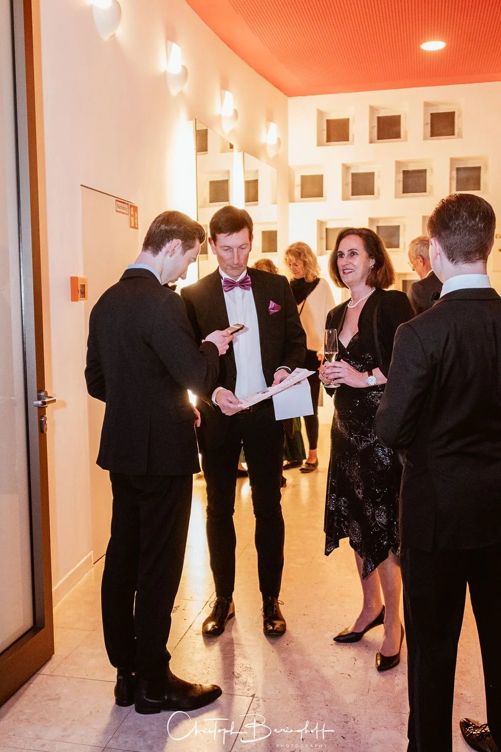 Vier elegante Personen in formeller Kleidung sprechen bei einer Veranstaltung, eine Frau hält ein Glas Sekt, zwei Männer tragen Anzüge, einer trägt eine Fliege, im Hintergrund weitere Gäste.