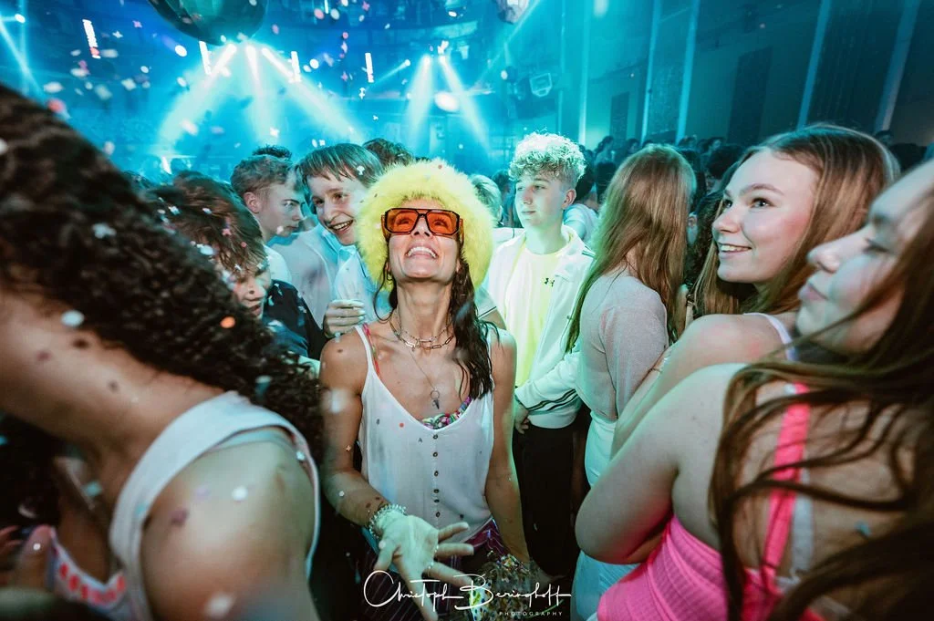 People are celebrating at a party with lights and fog effects; a woman wearing a yellow hat, large glasses, and sunglasses is at the center of the scene, surrounded by laughing teenagers.