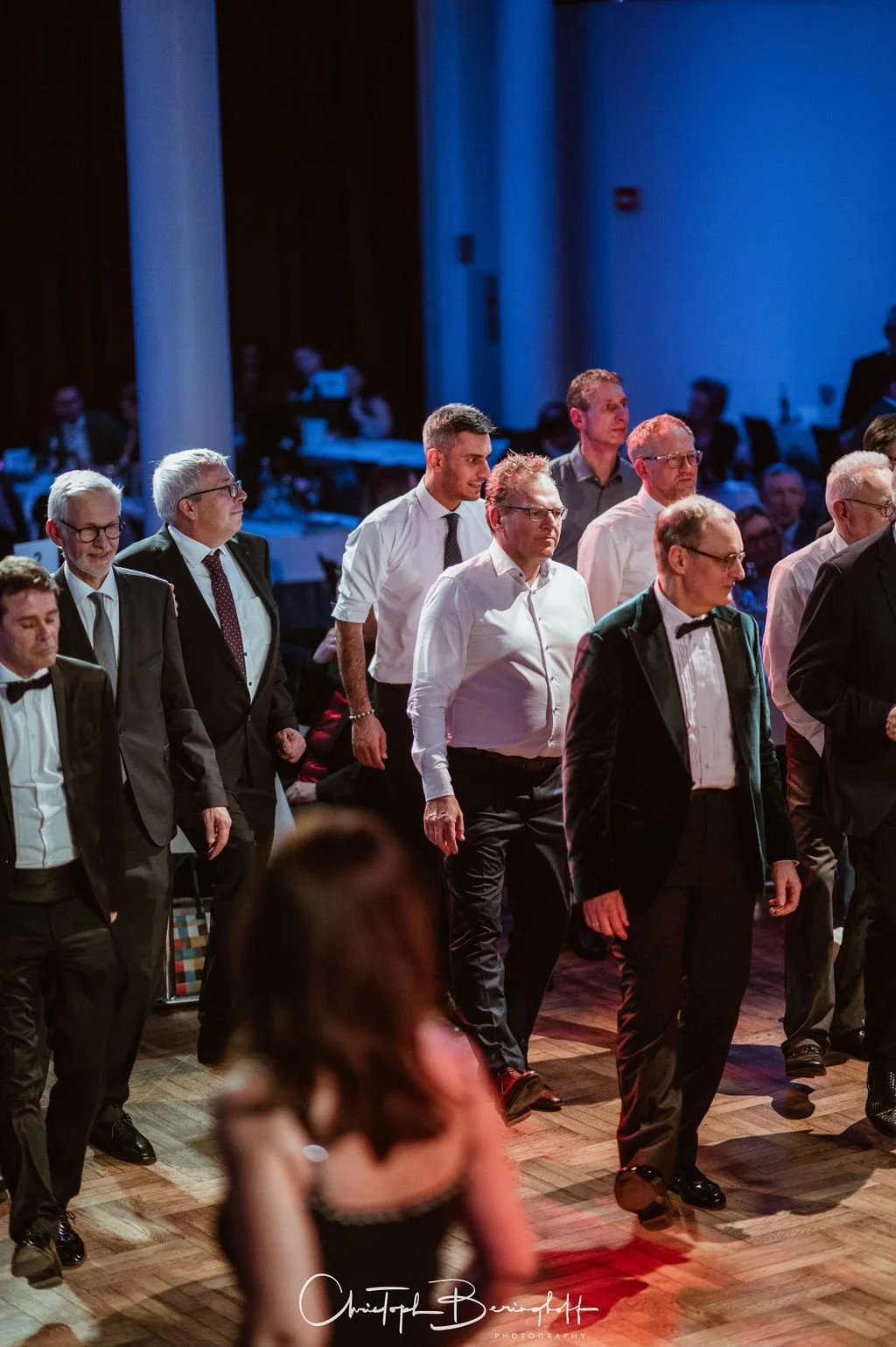 Mehrere Männer in formeller Kleidung, einige mit Anzügen und Krawatten, tanzen auf einer Bühne bei einer Gala-Veranstaltung. Im Hintergrund sitzen weitere Gäste an Tischen.