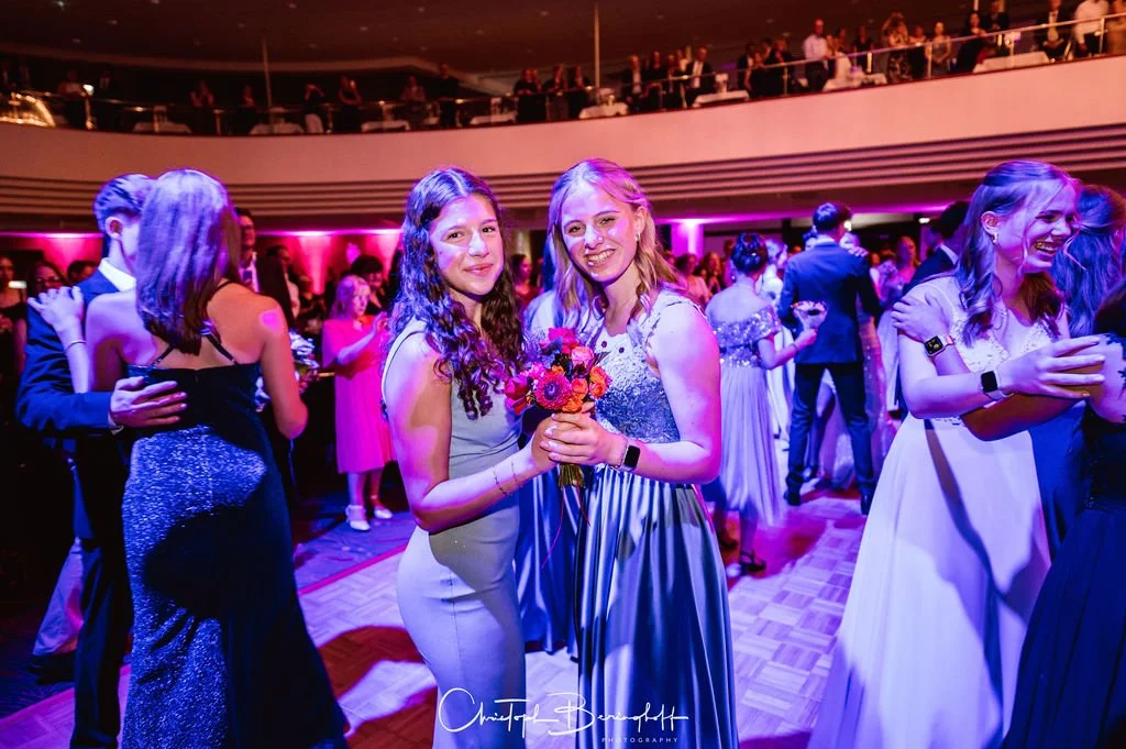 Zwei junge Frauen tanzen auf einer Party im Ballsaal, eine von ihnen hält einen Blumenstrauß, während andere Gäste im Hintergrund ebenfalls tanzen und feiern.