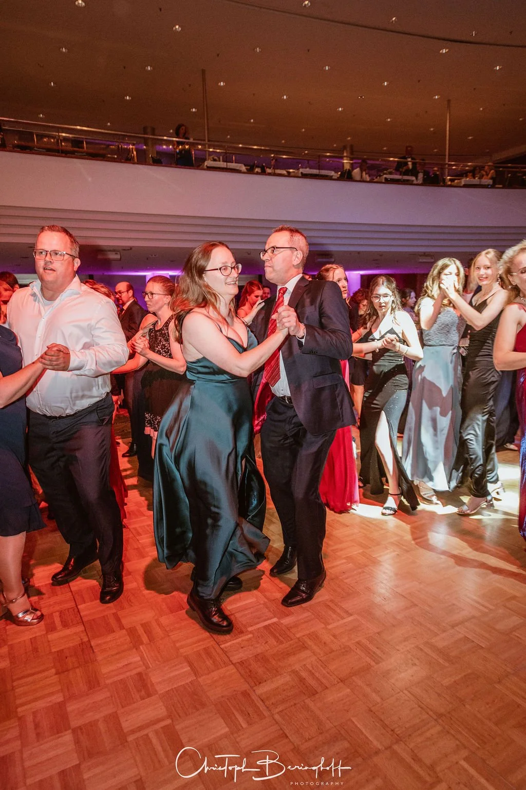 Menschen tanzen auf einer Feier im eleganten Abendkleid und Anzügen im hell erleuchteten Veranstaltungsraum.