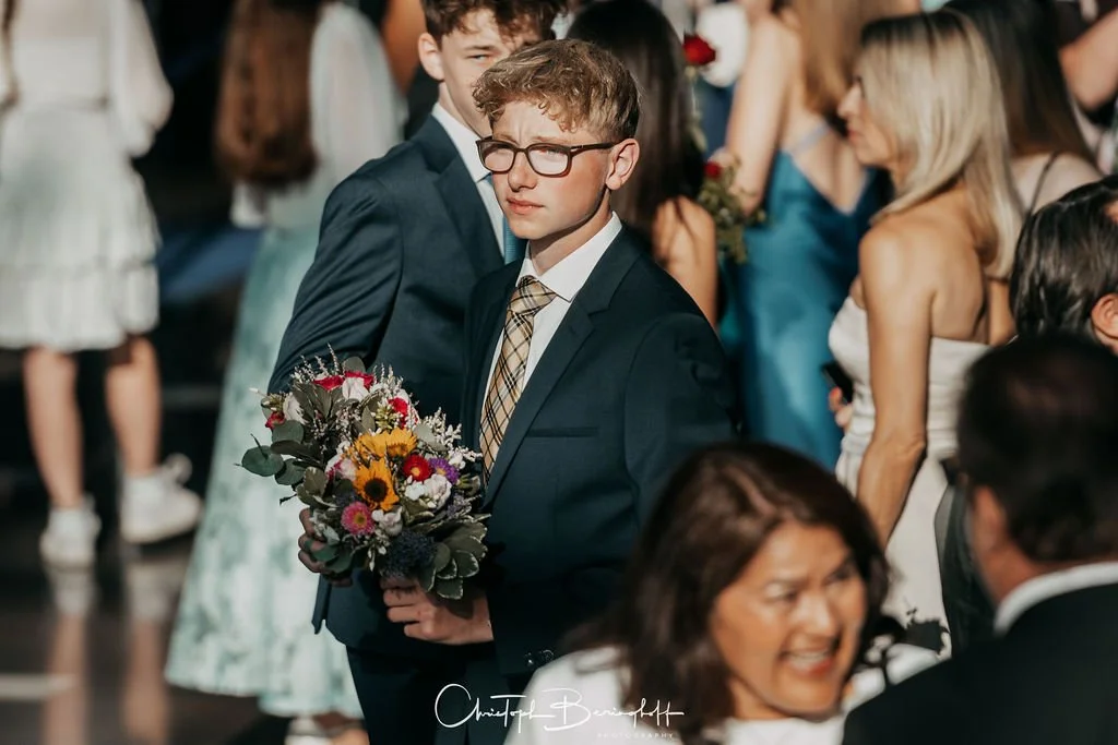 Junge Mann mit Brille und Anzug hält einen Blumenstrauß auf einer Feier, umgeben von Menschen in formeller Kleidung.