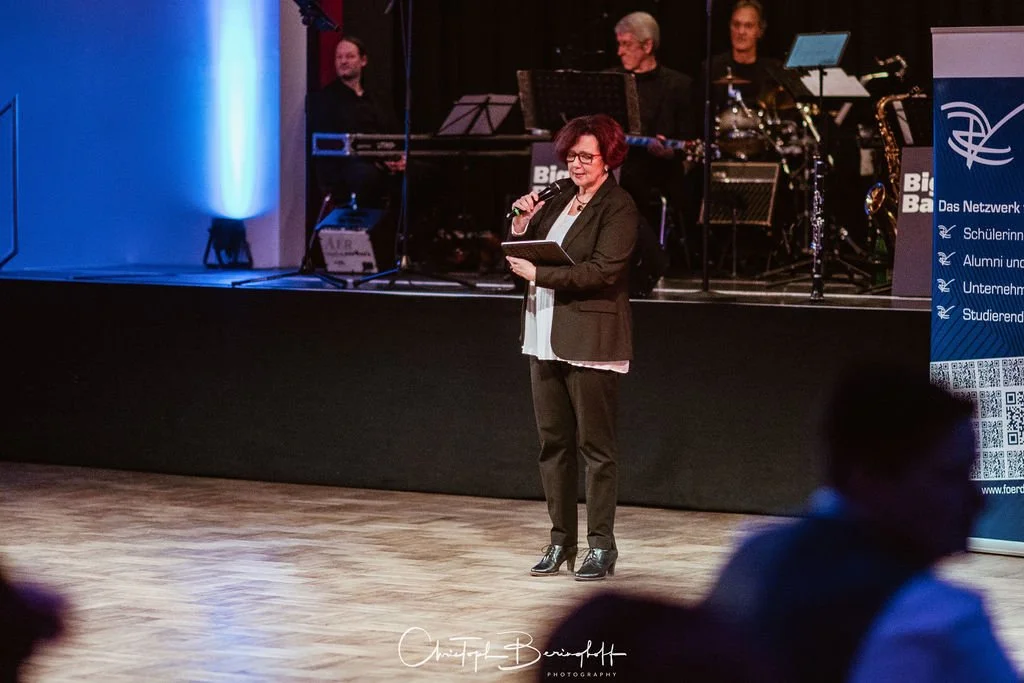 Eine Frau hält eine Rede oder Ansprache auf einer Bühne vor einem Mikrofon, im Hintergrund eine Band mit Musikern und Instrumenten, sowie ein blauer Banner mit Text.