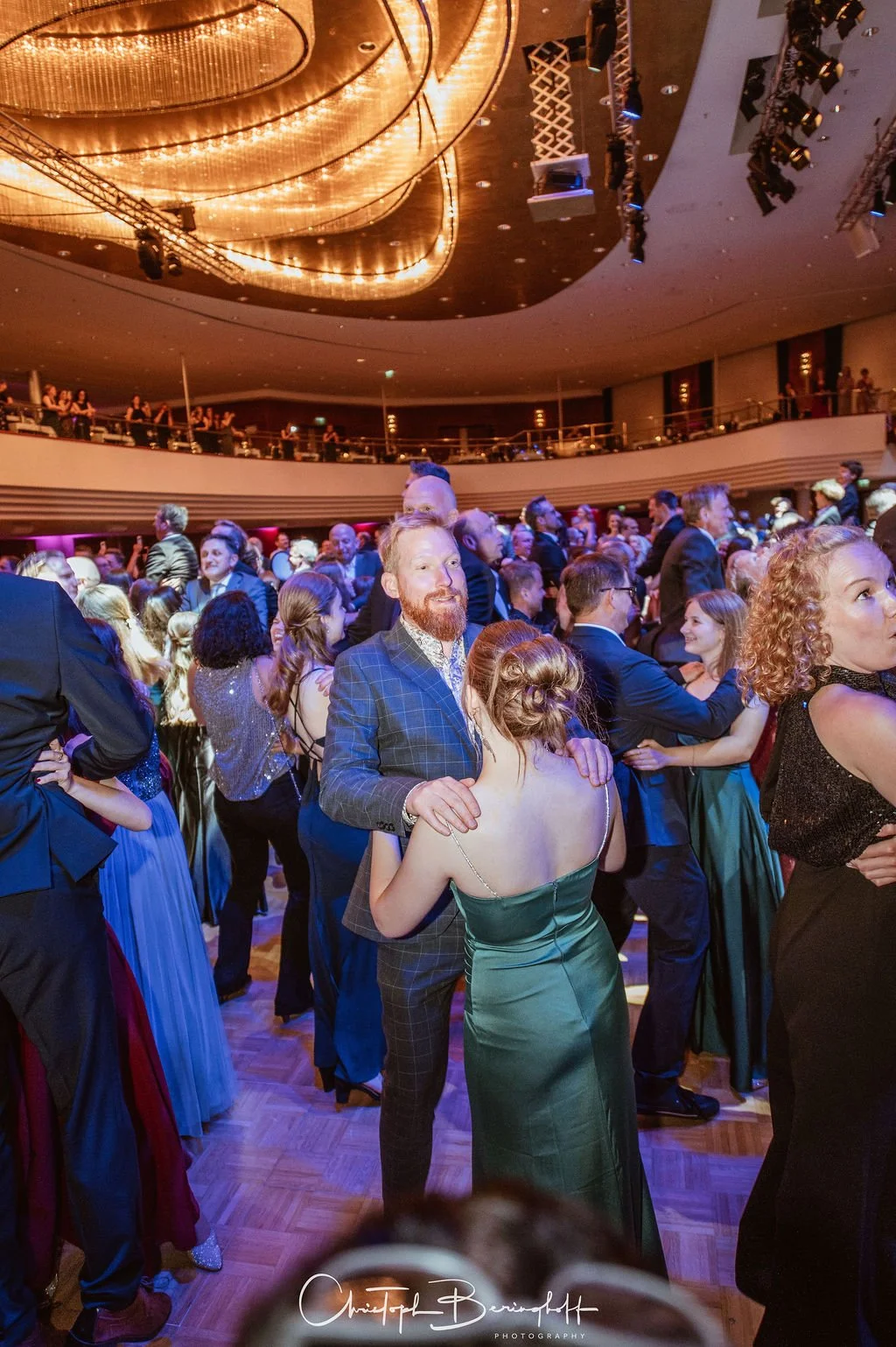 Menschen tanzen und feiern auf einer festlichen Veranstaltung im eleganten Saal mit Deckenleuchten und Lichtprojektionen.