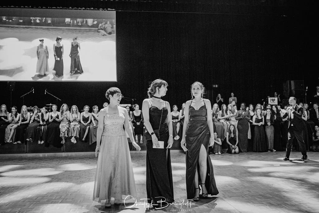 Schwarz-weiß-Fotografie einer Modenschau mit drei Frauen in eleganten Abendkleidern auf dem Laufsteg, im Hintergrund ein Publikum und ein großer Bildschirm