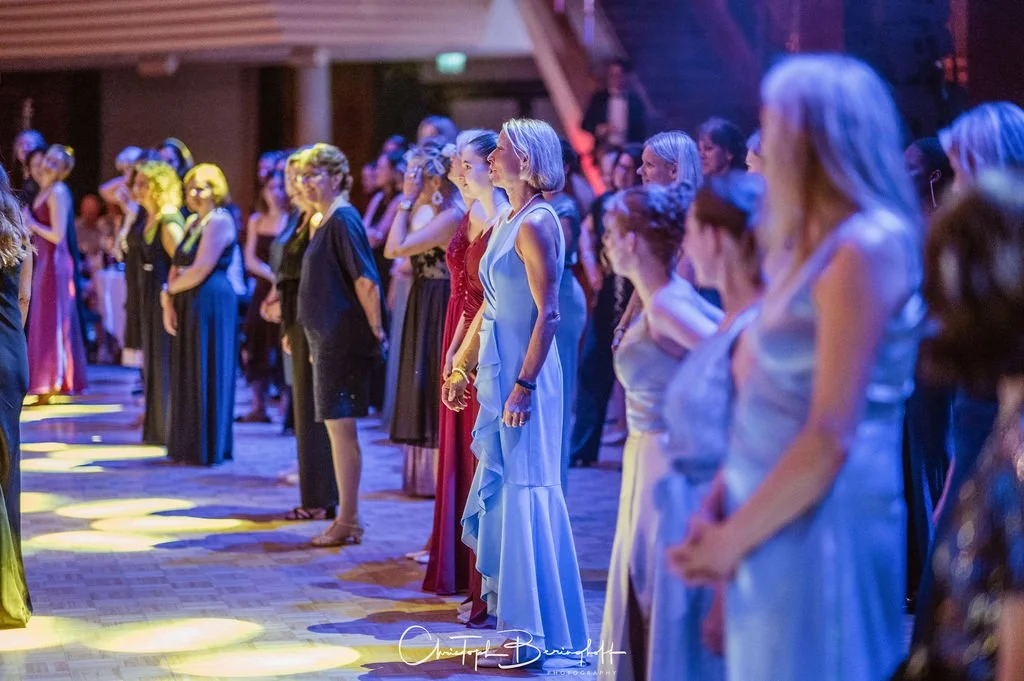 Frauengruppe beim Gala-Abend in eleganten Abendkleidern, stehender Empfang, festliche Atmosphäre, in einem Veranstaltungsraum mit lichtdurchfluteter Decke.