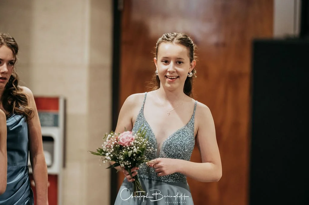 Eine junge Frau in einem silbernen Kleid hält einen Blumenstrauß und trägt Ohrringe und eine Halskette, während sie lächelt.