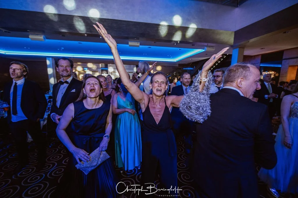 Junge und ältere Menschen feiern auf einer Party in formeller Kleidung, einige tanzen und lachen, im Hintergrund blaue Beleuchtung.