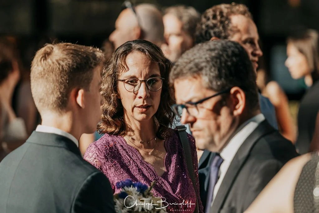 Frau mit Brille und braunen Haaren im Gespräch mit zwei Männern auf einer Gesellschaftsfeier.
