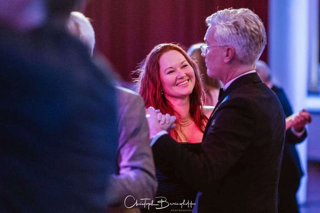 Ein Mann und eine Frau tanzen auf einer Party, die Frau lächelt glücklich, im Hintergrund weitere Gäste.