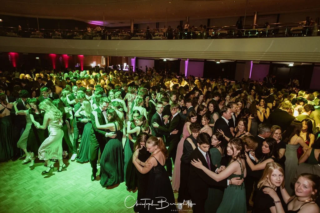 Eine große Gruppe von Menschen tanzt in einem festlichen Raum bei einer Veranstaltung oder einem Ball. Viele tragen elegante Kleidung.