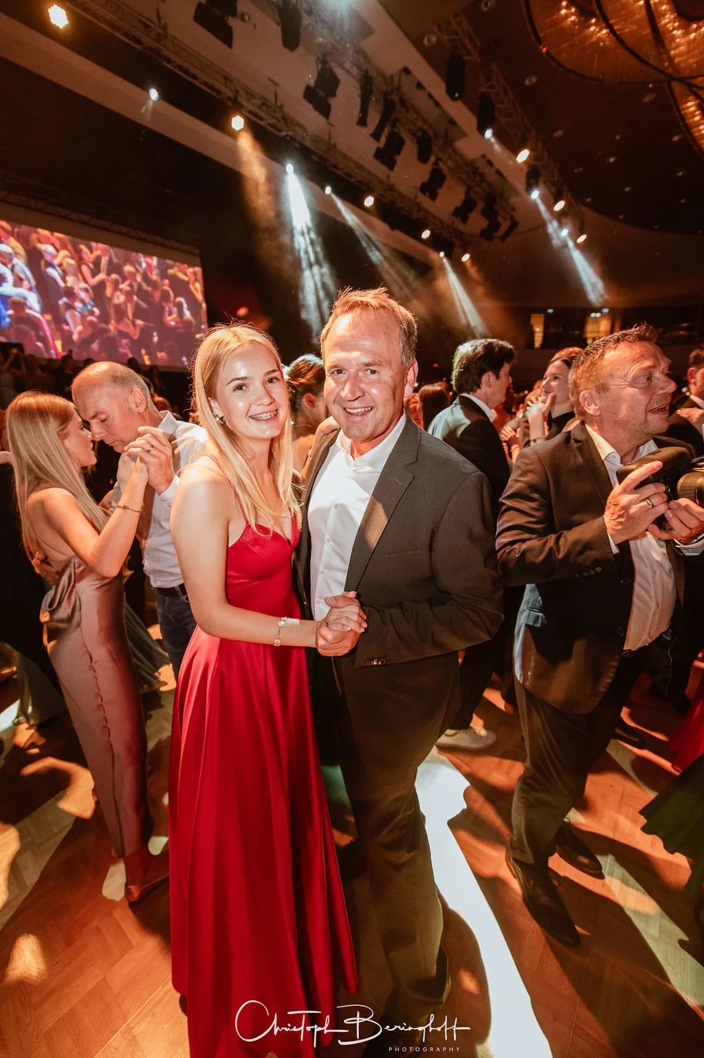 Ein Mann und eine Frau tanzen und lächeln auf einer festlichen Veranstaltung, umgeben von anderen Feiernden, mit bunten Lichteffekten und einer großen Leinwand im Hintergrund.