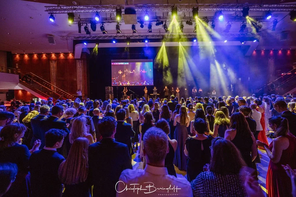 Menschen bei einer Veranstaltung im eleganten Stil, auf einer festlich beleuchteten Bühne mit Musikern und Schauspielern, in einem großen Saal mit rotem Akzent-Wänden.