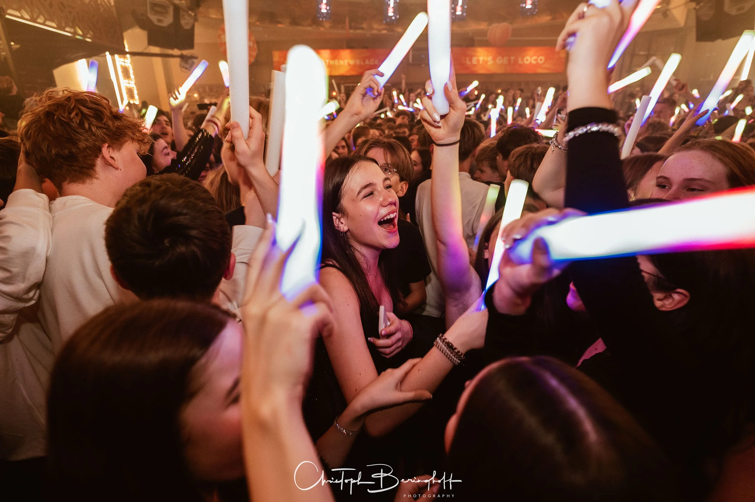 Eine Gruppe junger Menschen tanzt und feiert in einem dunklen, beleuchteten Raum mit leuchtenden Lightsticks, lachend und fröhlich.