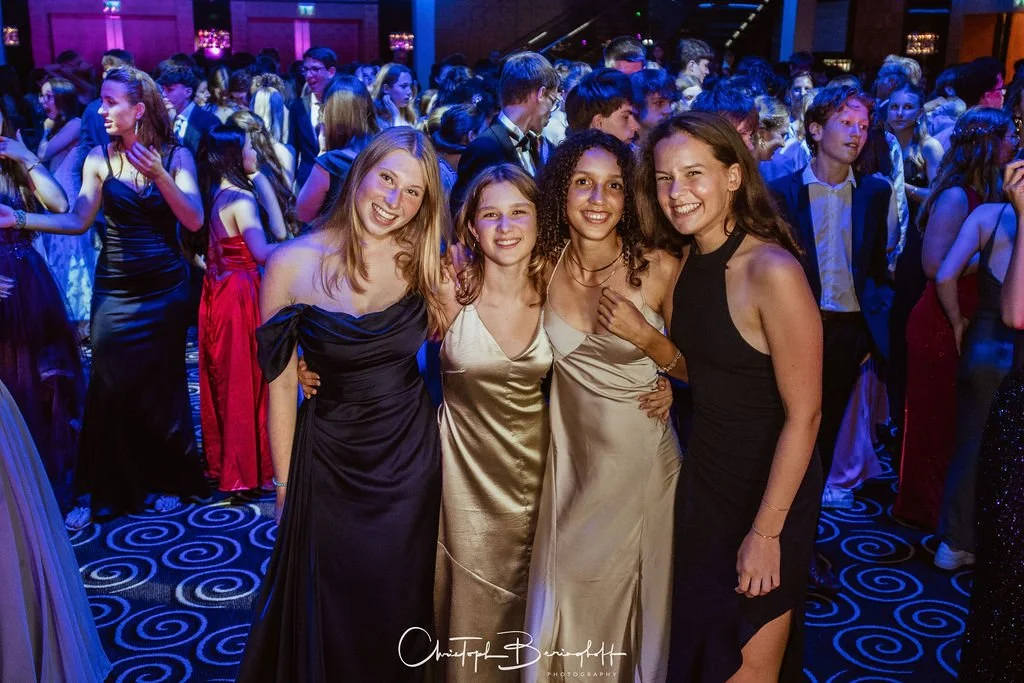 Vier junge Frauen in eleganten Kleidern posieren in einer festlichen Veranstaltungshalle, umgeben von vielen weiteren Gästen auf der Tanzfläche.