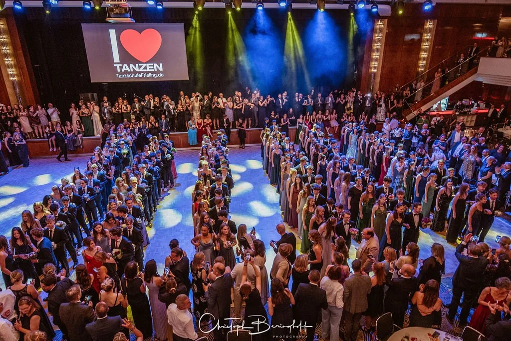 Tanzschule mit vielen Tanzpaaren, tanzen im großen Saal, eine große Menschenmenge, Bühnenbeleuchtung, LED-Leinwand mit 'Ich liebe Tanzen' in Deutsch.