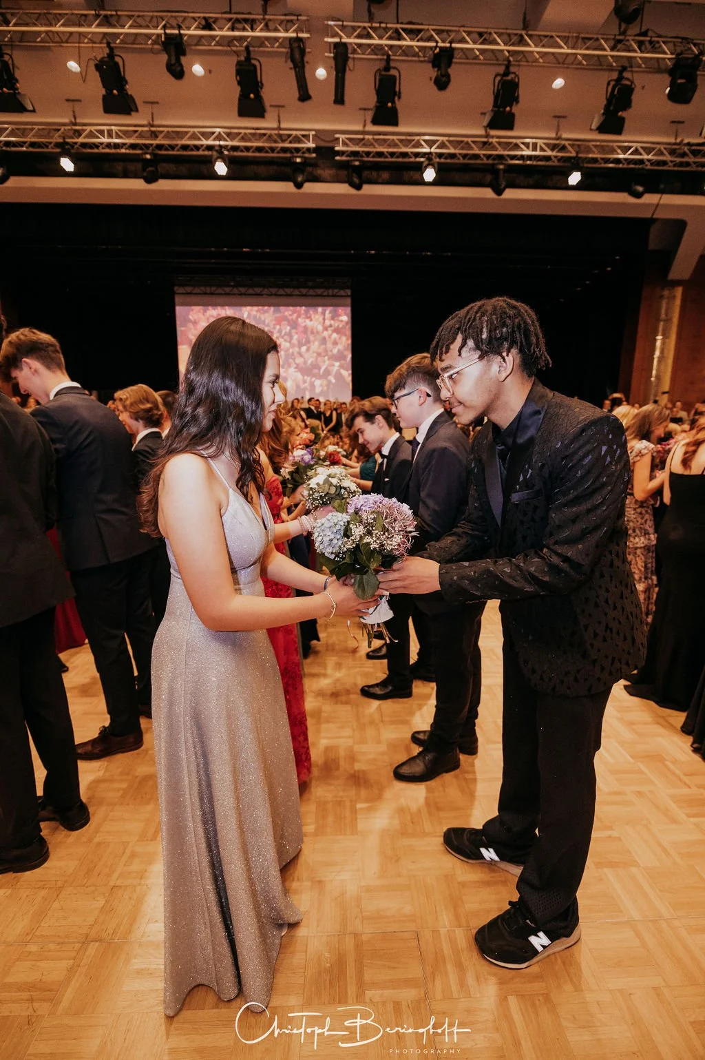 Ein junges Paar bei einer formellen Veranstaltung tauscht Blumen aus, umgeben von anderen elegant gekleideten Menschen in einem großen Saal mit Bühne und Beleuchtung.