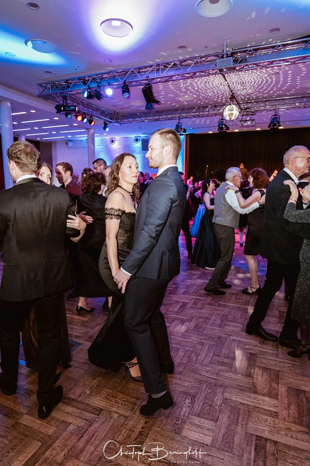 Menschen tanzen auf einer Party in formeller Kleidung im festlichen Raum, mit Tanzfläche, Disco-Kugel und bunten Lichtern.