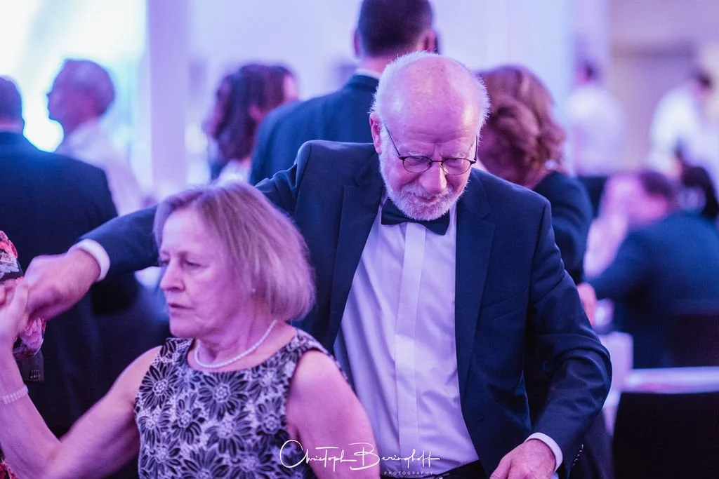 Ein älterer Mann in formeller Kleidung mit Brille und weißem Haar tanzt mit einer älteren Frau in einem eleganten Kleid auf einer festlichen Veranstaltung.
