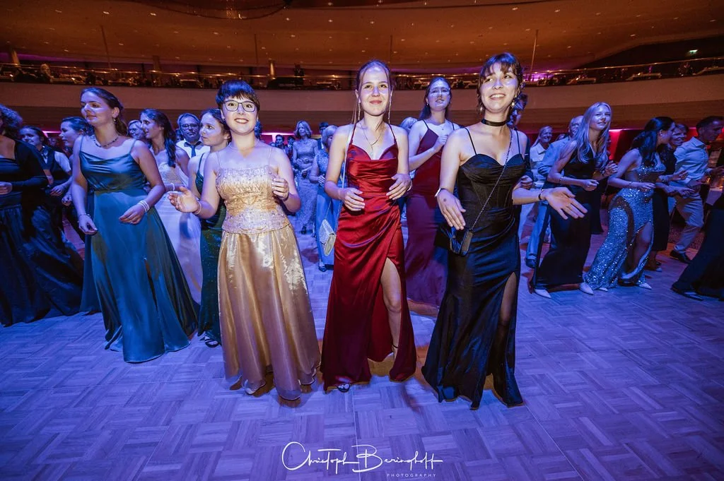 Junge Frauen in eleganten Abendkleidern tanzen auf einem Ballsaal. Die Stimmung ist festlich und fröhlich.