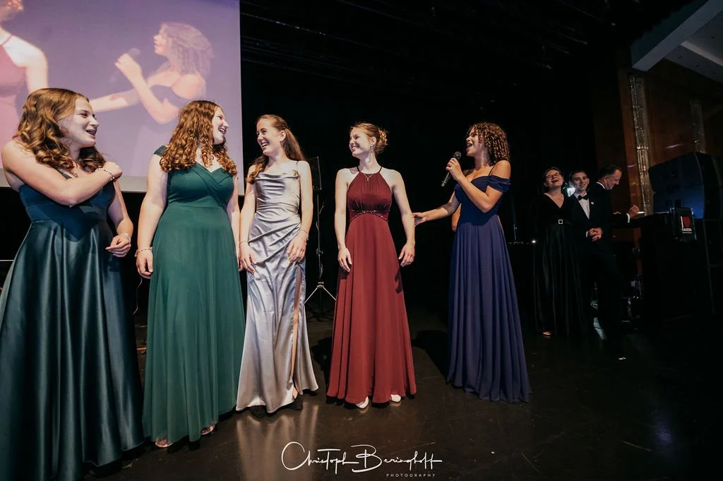 Sechs Frauen in langen, eleganten Abendkleidern stehen auf einer Bühne, eine spricht in ein Mikrofon, während die anderen lachen und zuschauen.