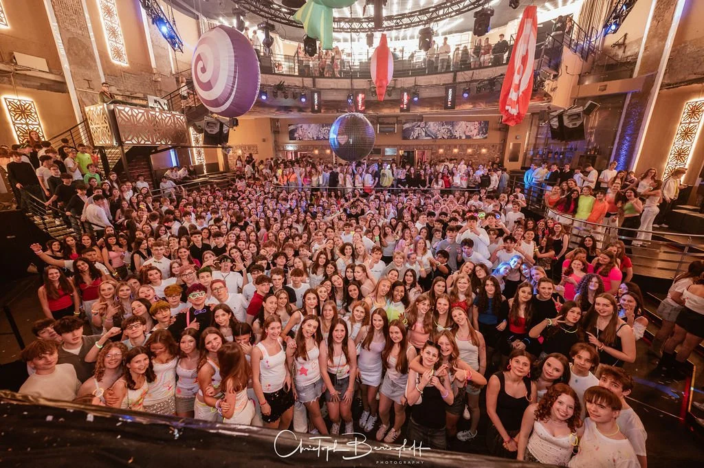 A large dance floor filled with young people at a club or party venue, with decorations hanging from the ceiling and several levels where more people can be seen watching.