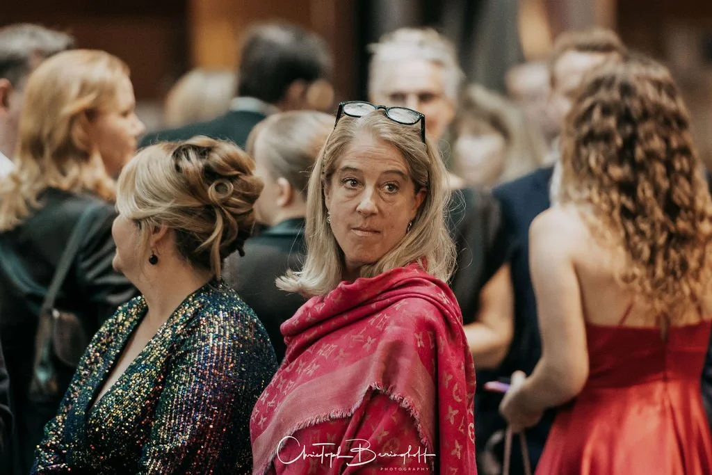 Eine Frau mit blondem Haar und Brille auf dem Kopf steht inmitten einer Gruppe von Menschen bei einer Veranstaltung.