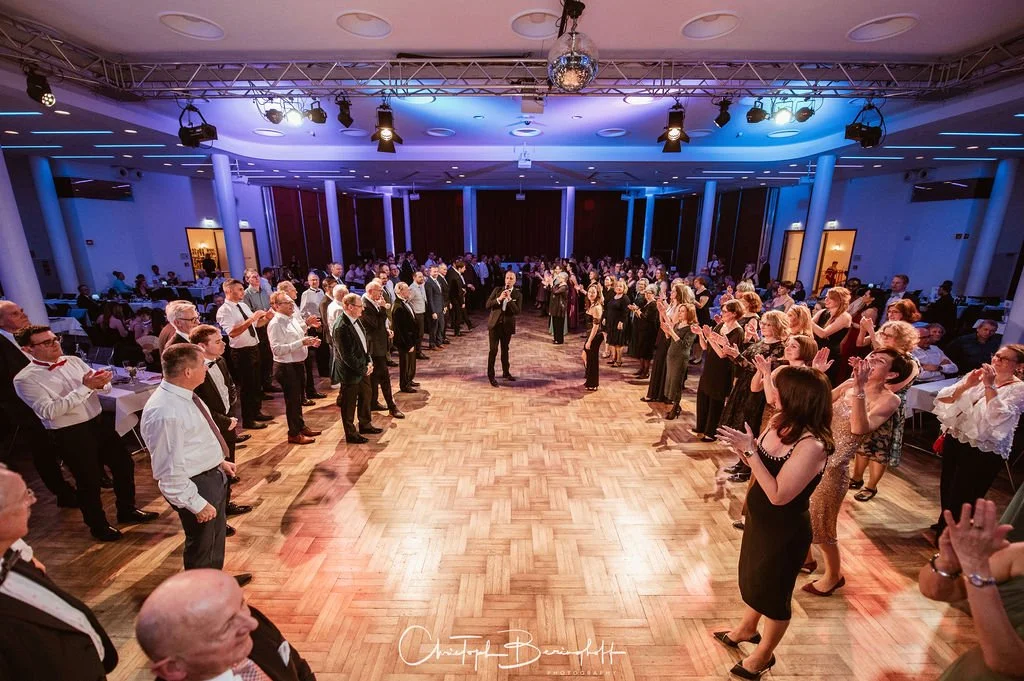 Menschen bei einer Tanzveranstaltung in einem Ballsaal, die in einer großen Gruppe im Kreis stehen.