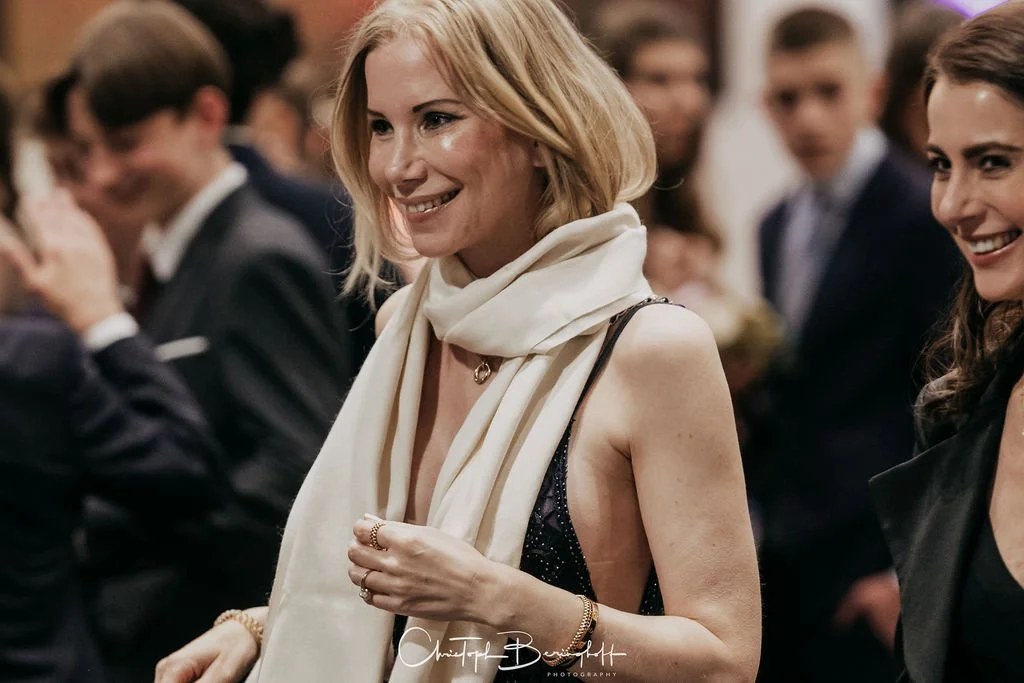Eine blonde Frau in einem schwarzen Kleid mit einer weißen Schärpe, lächelnd bei einer Veranstaltung, umgeben von anderen Menschen in formeller Kleidung.
