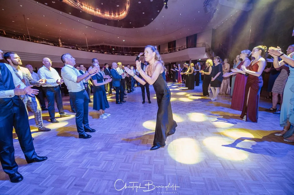 Tanzveranstaltung mit mehreren Menschen in einem Saal, der im Mittelpunkt einer Frau in schwarzem Kleid steht, die tanzt, während die anderen zuschauen und applaudieren.