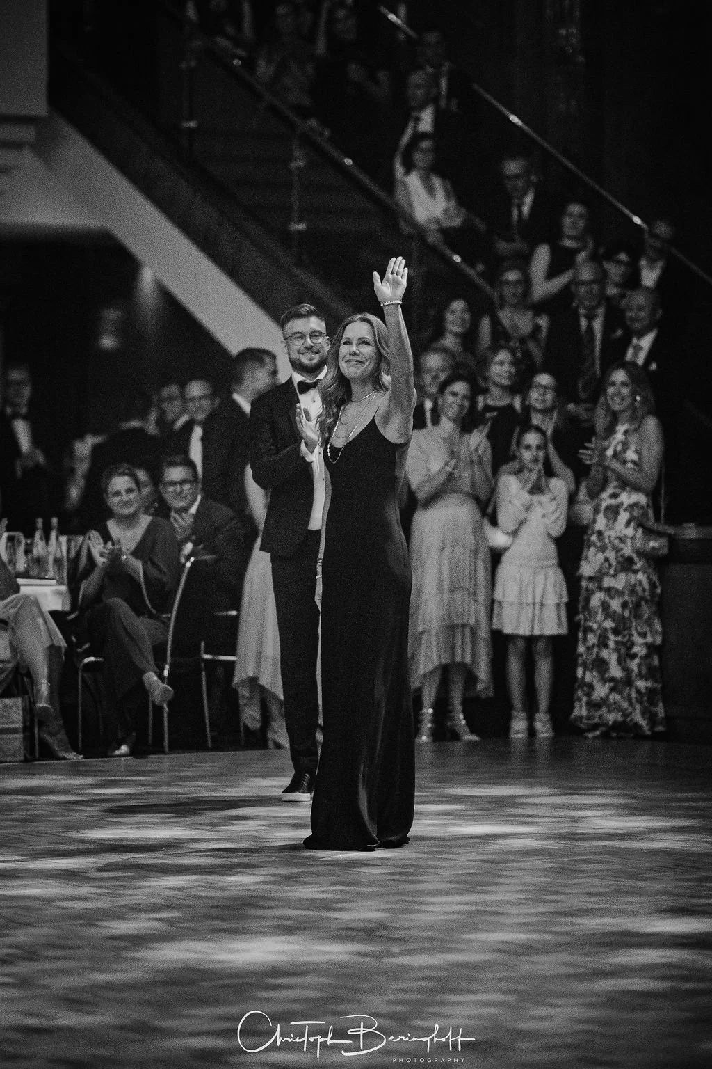 Ein elegantes Paar tanzt bei einer formellen Veranstaltung, umgeben von einem applaudierenden und lachenden Publikum, im Schwarz-Weiß-Foto aufgenommen.