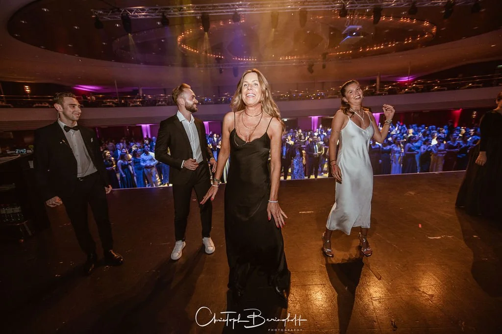 Menschen tanzen auf einer Party im großen Saal, bunt beleuchtete Tanzfläche, festliche Kleidung, Stimmung fröhlich.