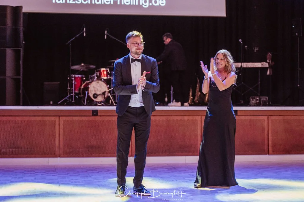 Ein Mann in Smoking und eine Frau in einem schwarzen Abendkleid stehen auf einer Bühne und klatschen, im Hintergrund ist eine Band mit Schlagzeug und einem Keyboard zu sehen.