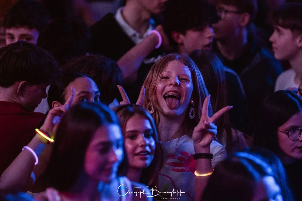 People at a party, with a woman at the center, playing and sticking out her tongue, surrounded by others, in a dark, colorful setting.