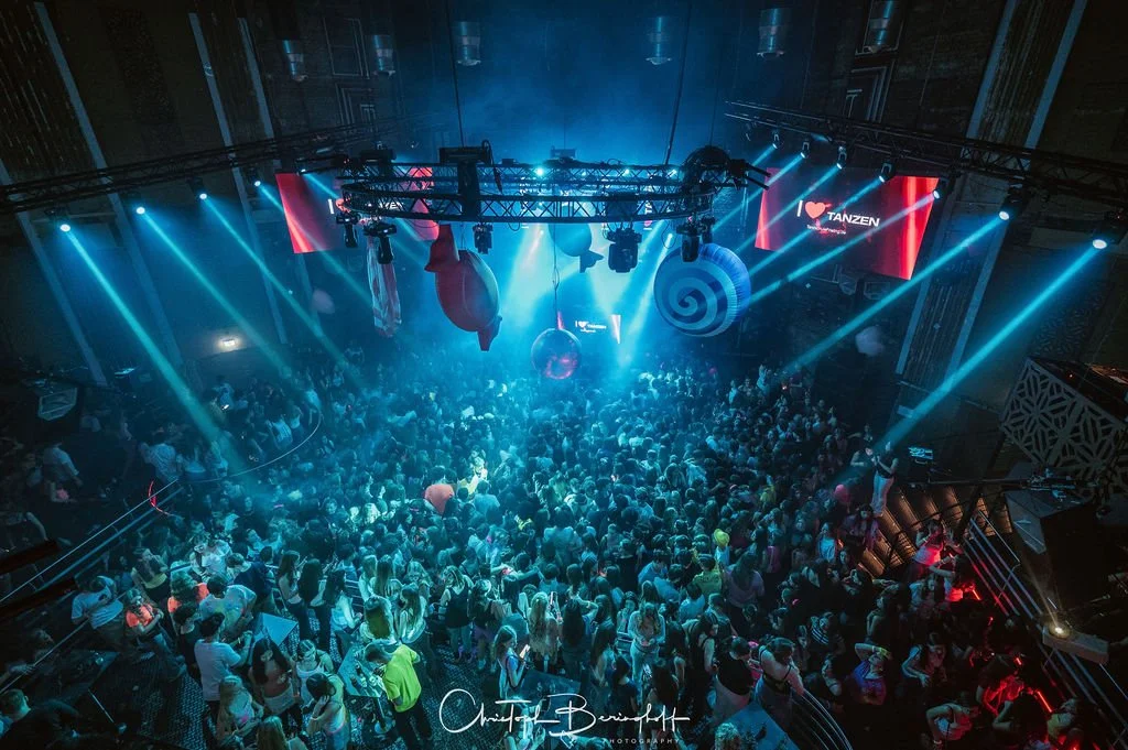 A large club or event venue filled with people, colorful lights, and a stage where a performance is taking place, with digital screens in the background.